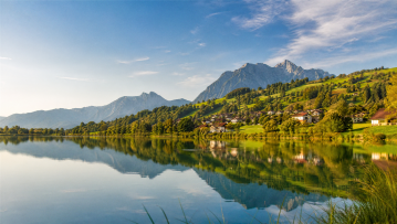 Idyllische Landschaft in Waltenhofen mit einem See, an dessen Ufer sich Häuser an einen grünen Hügel schmiegen, dahinter ragen Berge in den blauen Him
