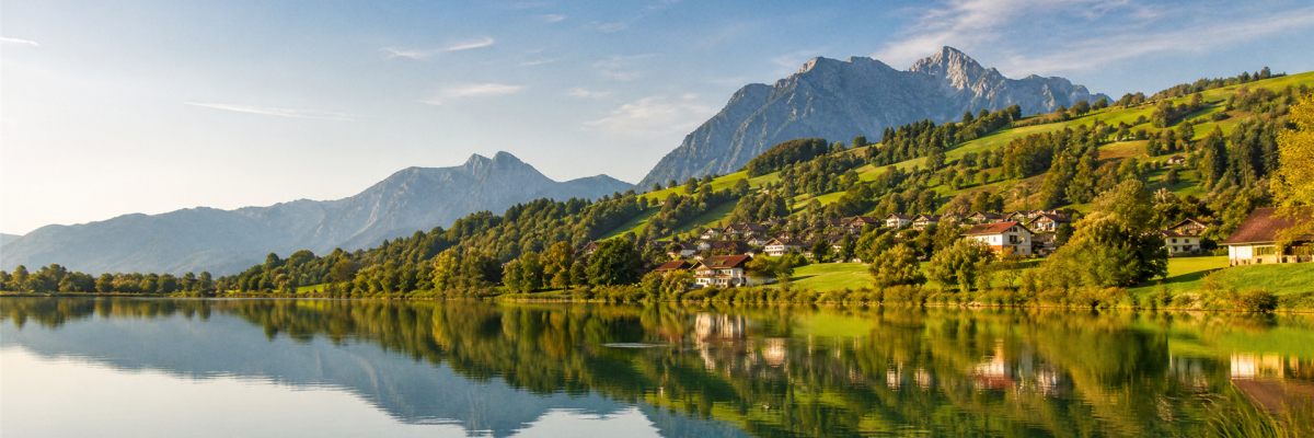 Idyllische Landschaft in Waltenhofen mit einem See, an dessen Ufer sich Häuser an einen grünen Hügel schmiegen, dahinter ragen Berge in den blauen Him