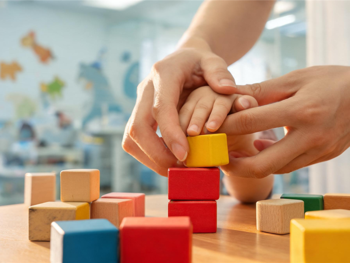 Eine Handtherapeutin führt sanft die Hand eines Kleinkindes beim spielerischen Stapeln von bunten Holzklötzen in einer hellen Praxis.
